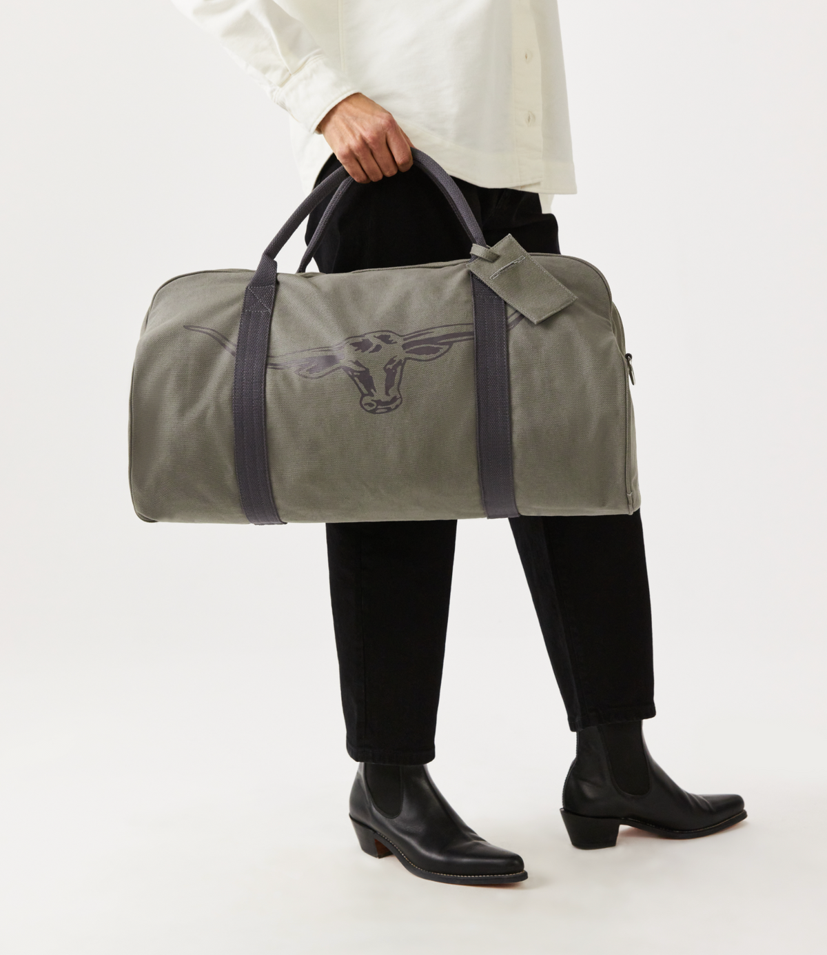 A person wearing a white shirt and black pants holds the gray RM Williams Nanga Canvas Bag with a cow design. The cotton canvas material pairs well with their black boots, all set against a plain white backdrop.
