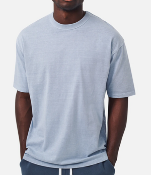 A person wears the Industrie The Del Sur Tee, an oversized light blue T-shirt made of 100% cotton, paired with dark blue pants. They stand against a plain white background with hands in pockets. The casual tee is simple and free from visible logos or designs.