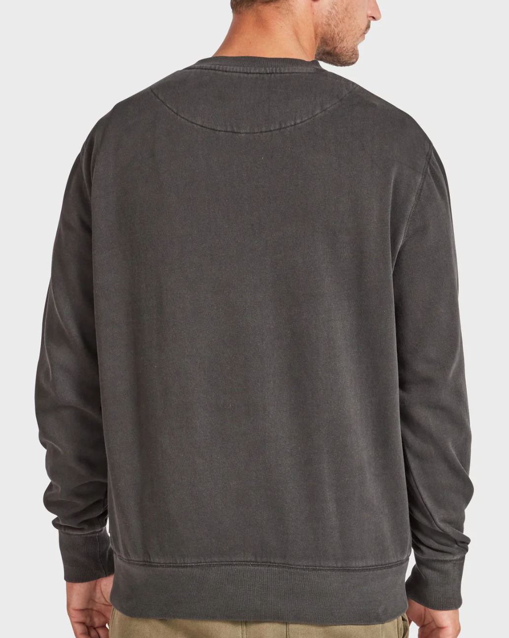 A man is seen from the back against a plain white background, wearing the Academy Brand Crew Sweat in relaxed fit, long sleeves, dark gray, paired with beige pants.