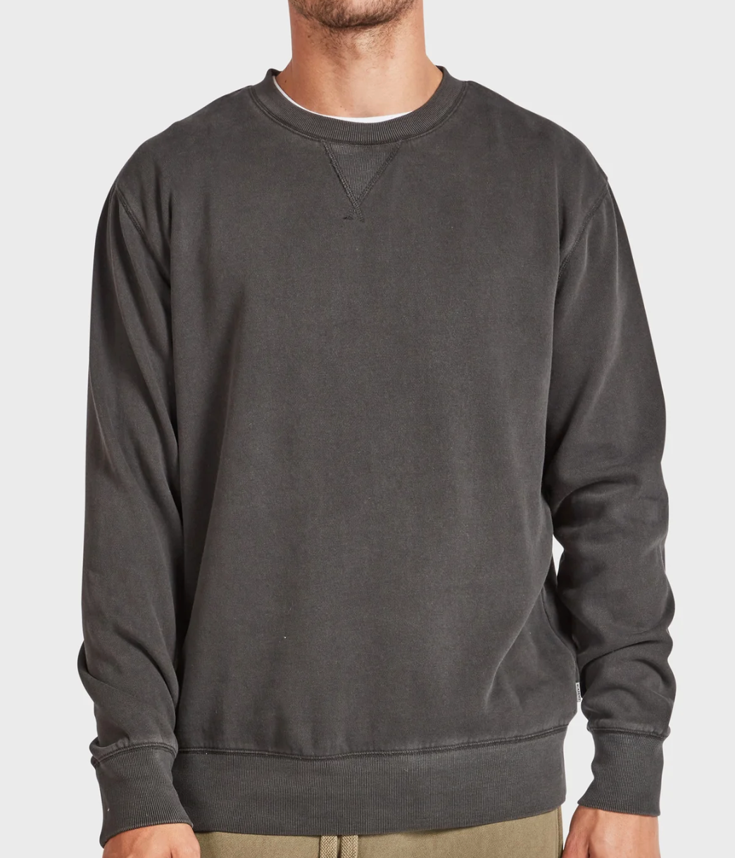 A person is wearing the Academy Brand Crew Sweat, a plain, long-sleeve, dark gray brushed fleece crewneck sweatshirt with khaki pants. The image captures their upper body against a light background.