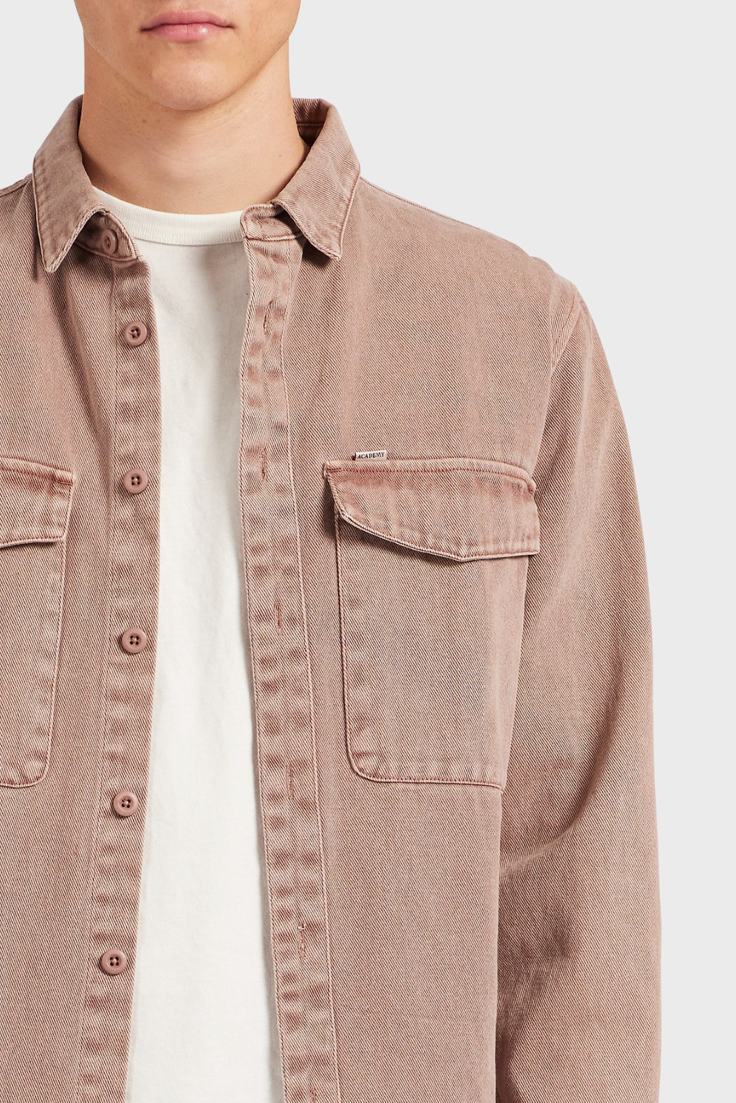 A person wears the Academy Brand Essential Overshirt in dusty pink 100% cotton, styled open over a light beige tee. The overshirt features two chest pockets and is shown from shoulders to mid-torso against a plain background.