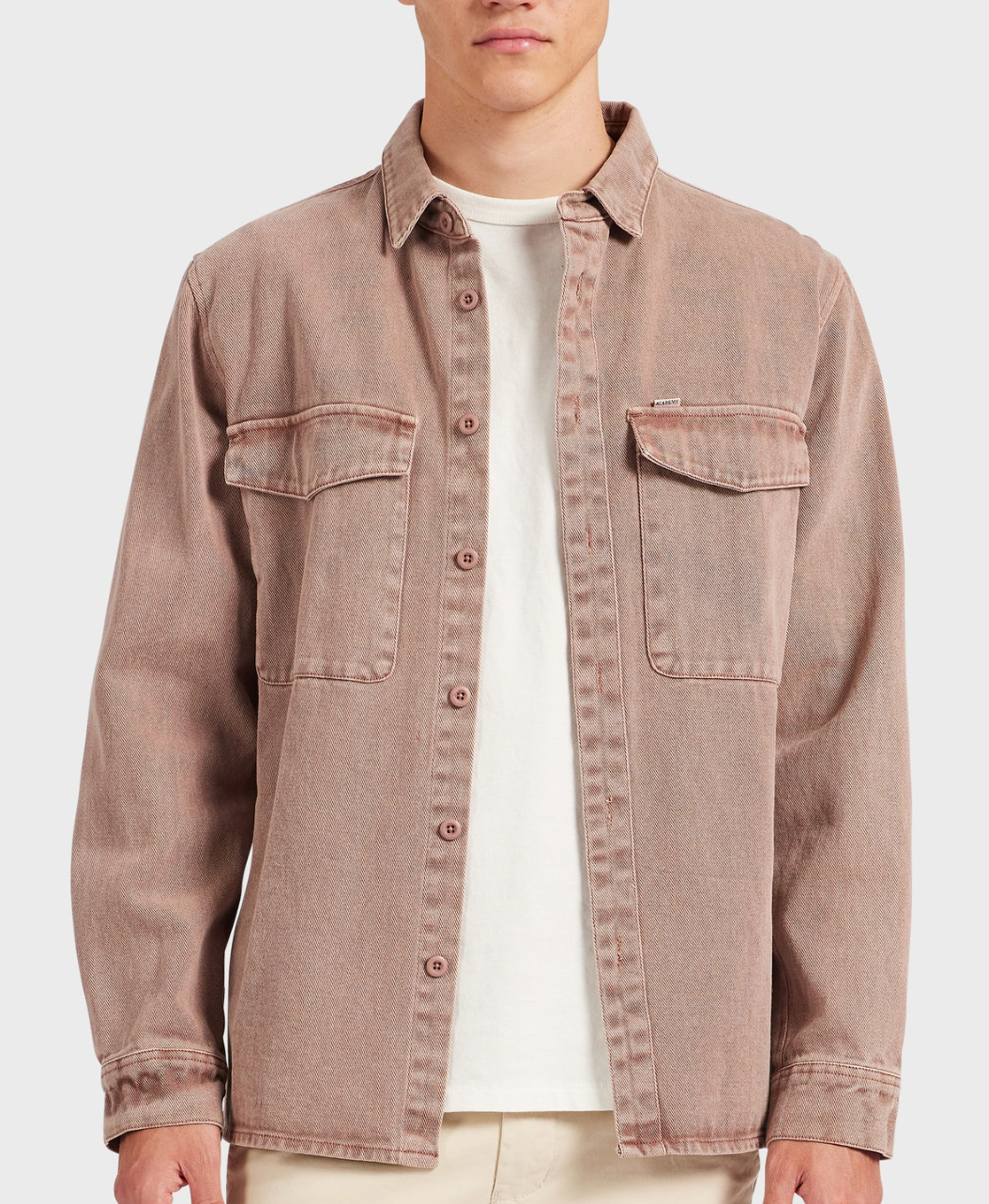 A person wears the Academy Brand Essential Overshirt in light brown 100% cotton with chest pockets, layered over a white t-shirt and beige pants, standing against a plain light background.