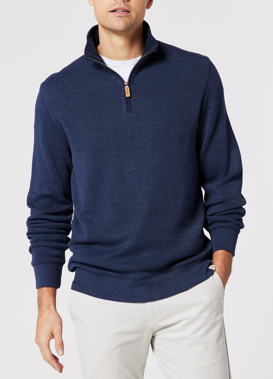 A man wears the Blazer Heather Half Zip Sweat, a navy peached-finish sweater with a half zip, over a white shirt and light pants. He stands against a plain light background with his left hand by his side and right arm slightly bent.
