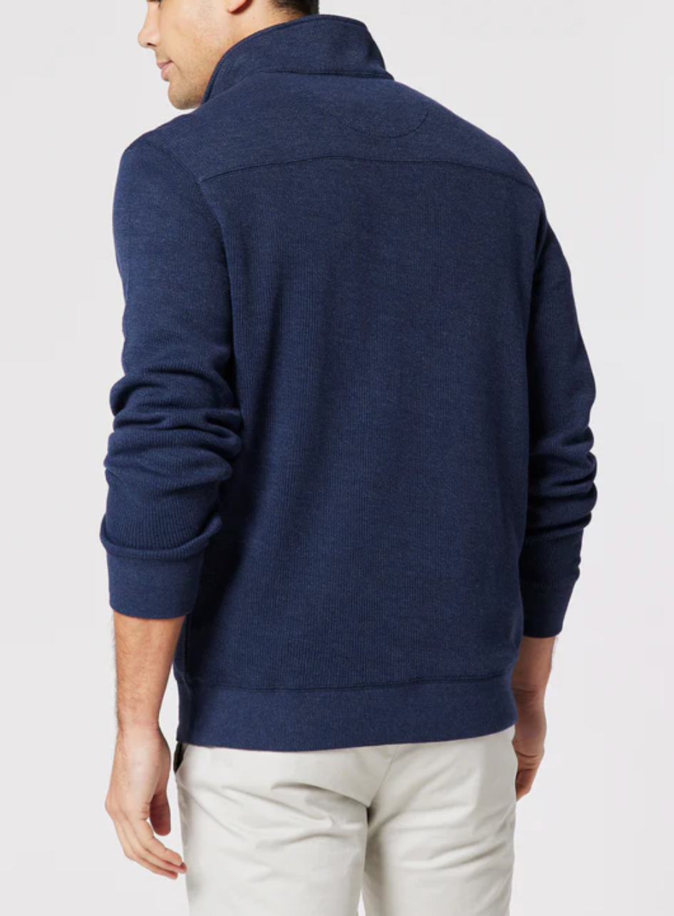 A man is seen from the back wearing the Blazer Heather Half Zip Sweat, a dark blue, long-sleeve collared sweater with a peached finish, paired with light pants, standing against a plain light background.