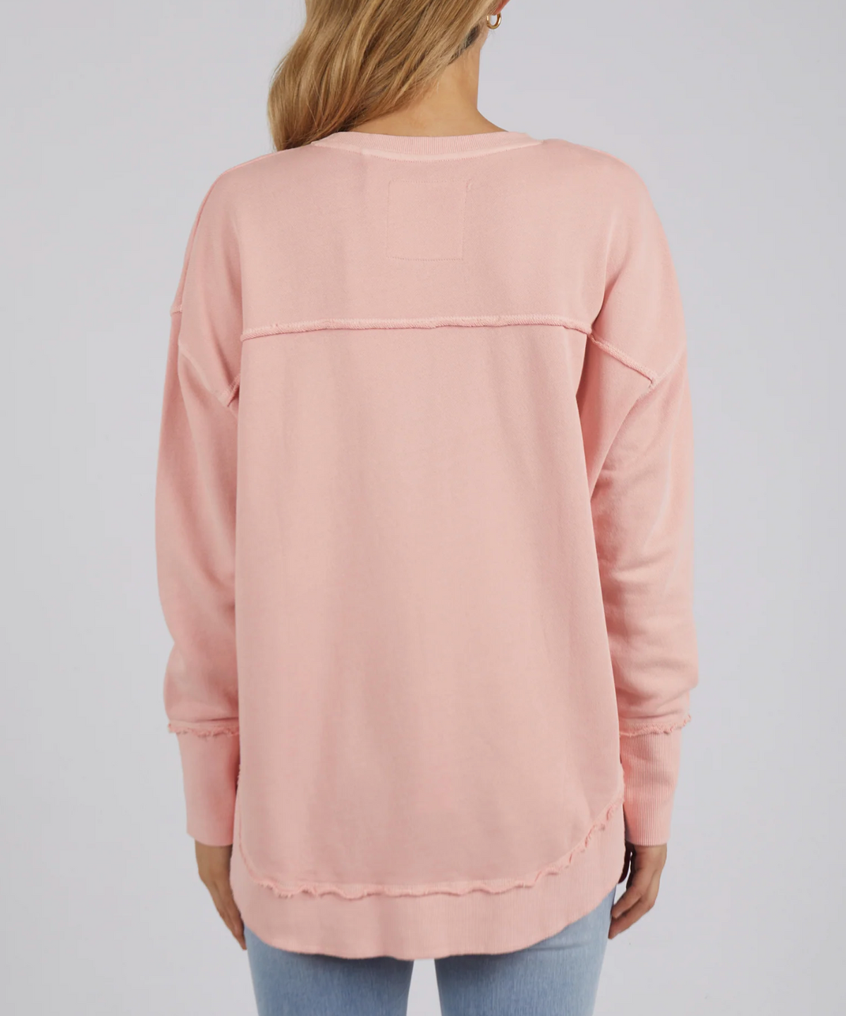 The rear view shows a person with long hair wearing the Foxwood Simplified Crew, an oversized light pink cotton jumper with visible seam detail across the back, paired with light blue jeans, set against a plain white background.