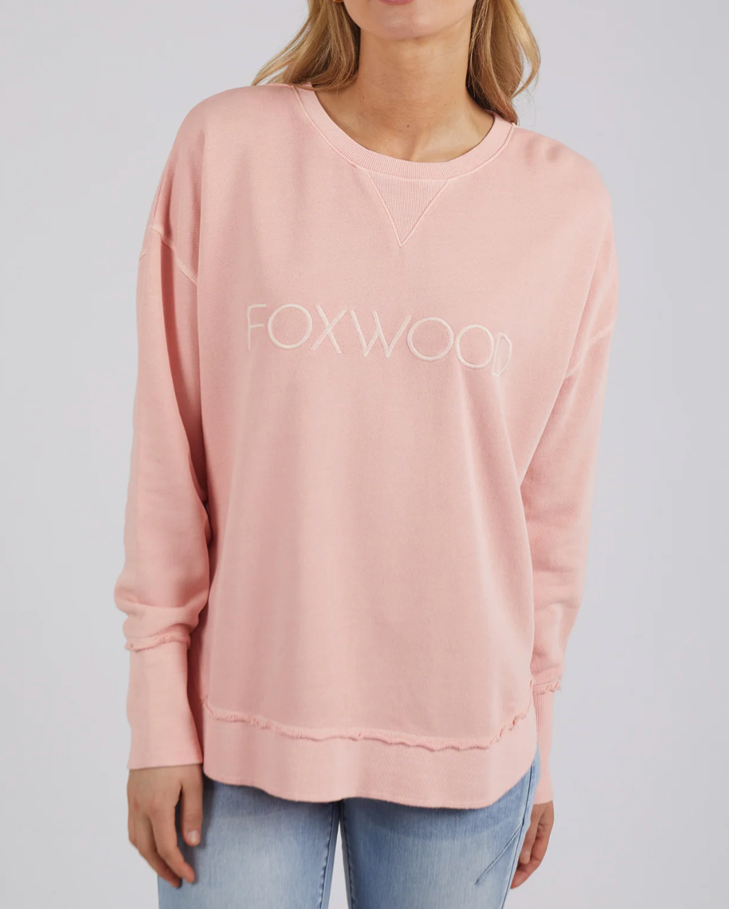 A person wears the Foxwood Simplified Crew, a light pink cotton jumper featuring "FOXWOOD" in white on the front. Its relaxed fit pairs effortlessly with light blue jeans against a white background, forming a serene and stylish look.