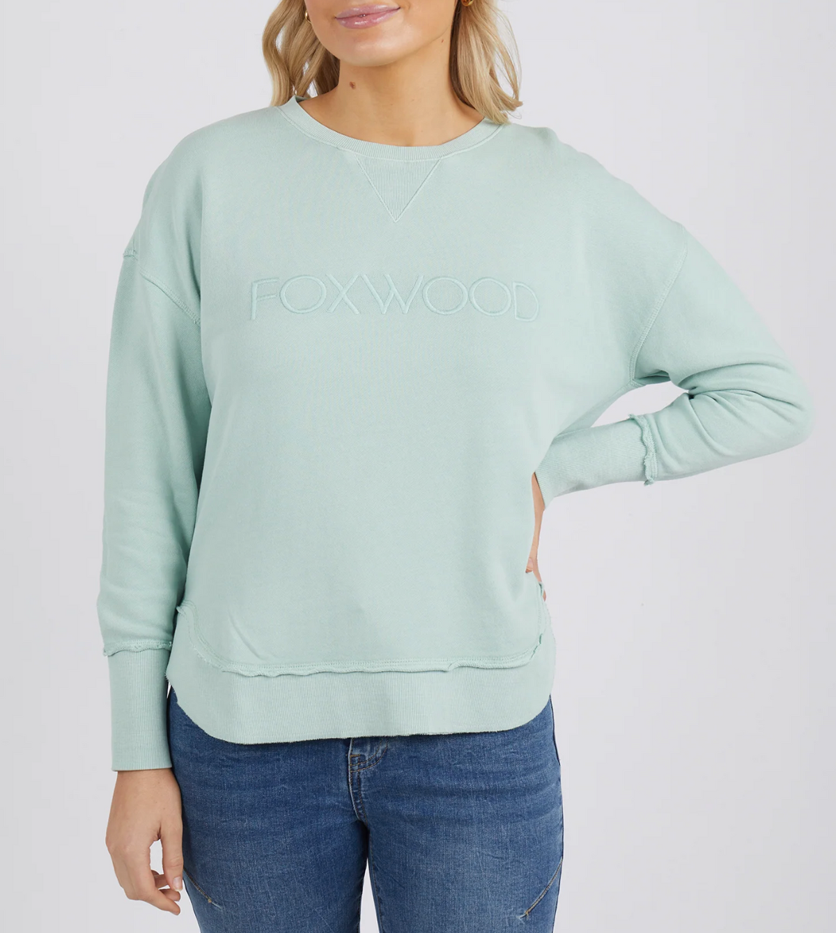 Someone is wearing the light green Foxwood Simplified Crew with "FOXWOOD" embroidered on the front, paired with blue jeans. Their left hand is on their hip against a plain white background.