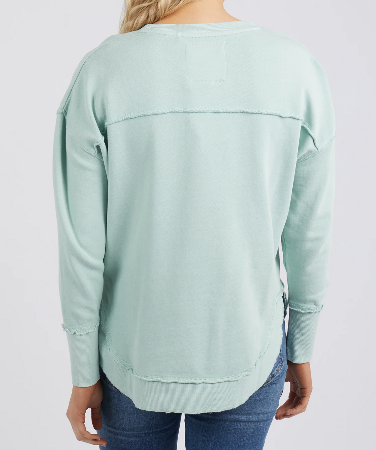 A person with long blonde hair, viewed from behind, wears a light turquoise Foxwood Simplified Crew sweatshirt made of 100% cotton and blue jeans. The relaxed-fit sweatshirt features visible seams, a slightly frayed hem, and subtle Foxwood embroidery.