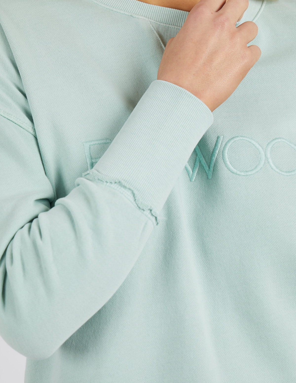 A person in the Foxwood Simplified Crew, a light green 100% cotton sweatshirt with subtle embroidered text, touches their neck with their right hand. The focus is on the sleeve's slightly frayed edges, and the background is not visible.