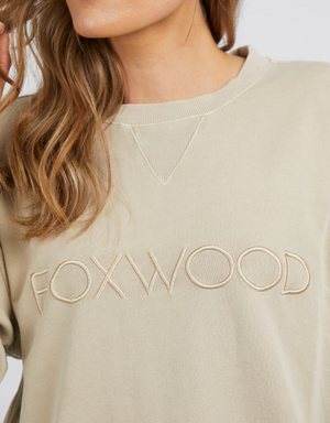 A woman with wavy light brown hair wears a beige Foxwood Simplified Crew jumper with "FOXWOOD" in large capital letters. Seen from the shoulders up, she is facing slightly to the side.