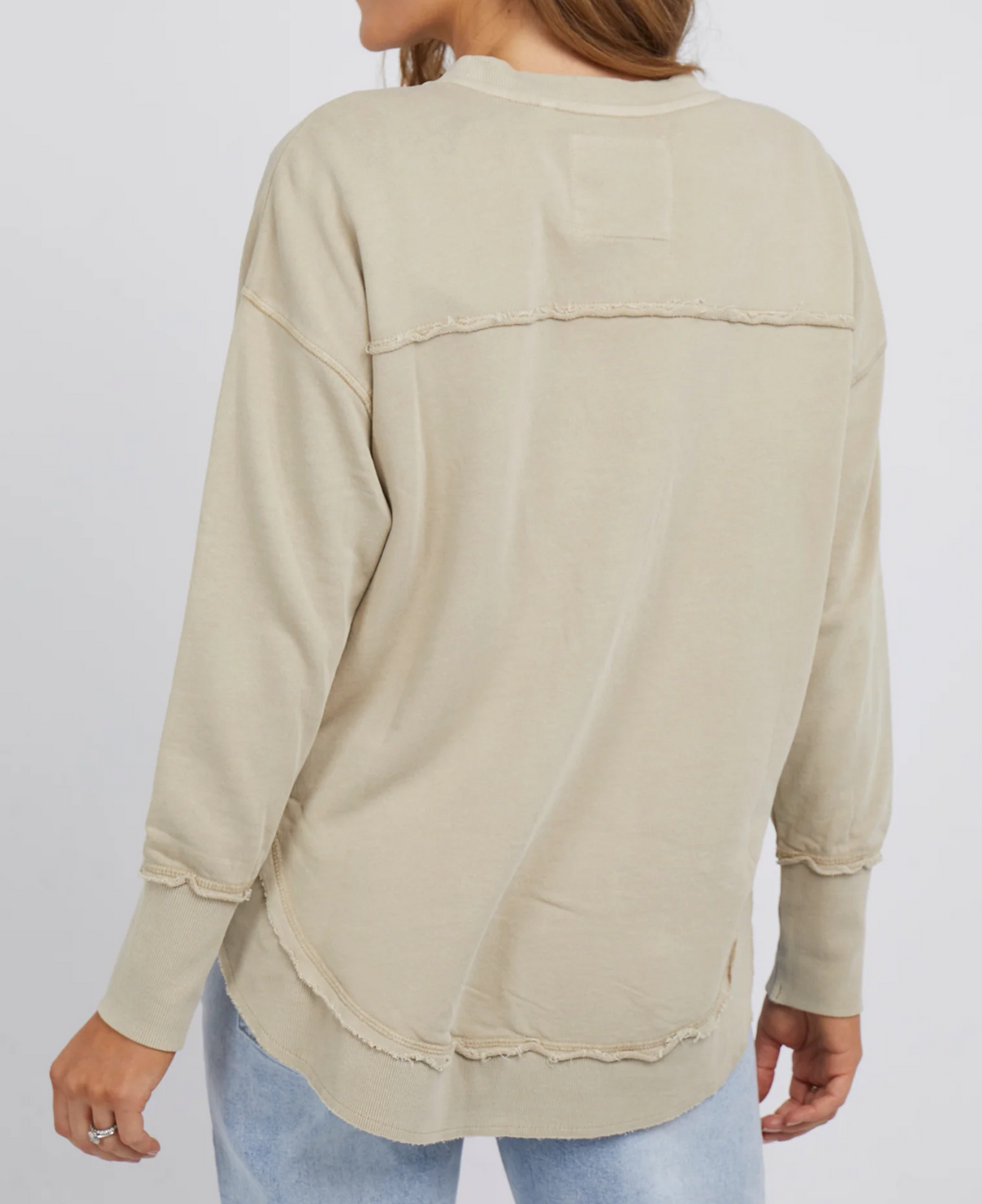 Rear view of someone in a beige Foxwood Simplified Crew jumper with frayed edges and blue jeans. The cotton jumper has unfinished seams across the back and hem. They stand against a plain white background.