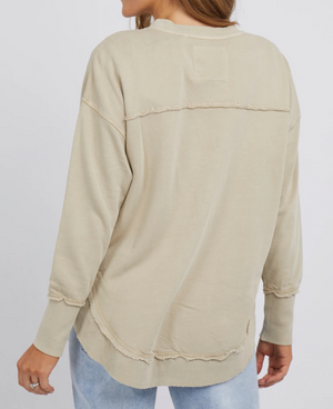 Rear view of someone in a beige Foxwood Simplified Crew jumper with frayed edges and blue jeans. The cotton jumper has unfinished seams across the back and hem. They stand against a plain white background.