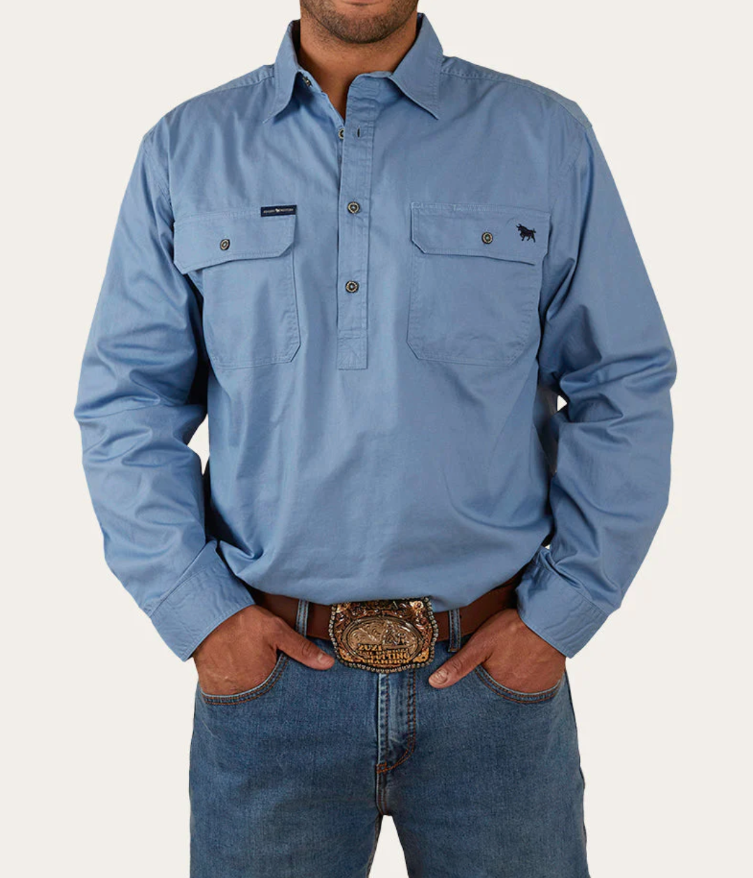 Dressed in a Ringers Western King River Half Button Shirt, 100% cotton, light blue with two chest pockets and an embroidered dog logo, paired with blue jeans and a large belt buckle, the style pops against the plain white background.