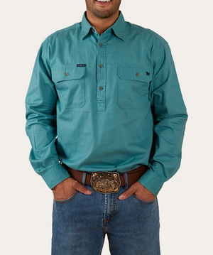 A man wearing a long-sleeved Ringers Western King River Half Button Shirt in teal, featuring two chest pockets, stands with his hands in the pockets of his blue jeans. He is also sporting a decorative belt buckle and is smiling. The 100% cotton shirt perfectly complements the plain and light-colored background.