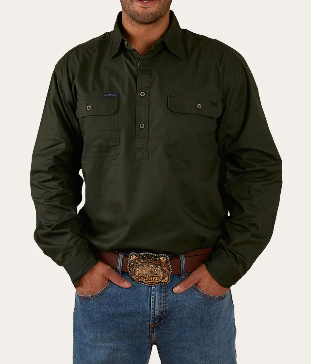 A person wearing a dark green Ringers Western King River Half Button Shirt, made of 100% cotton and featuring two front pockets, paired with blue jeans and a brown belt with an ornate buckle, is standing with their hands in their pockets against a plain, light-colored background.