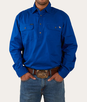 A person wears a bright blue Ringers Western King River Half Button Shirt, made from comfortable cotton, featuring two chest pockets along with jeans. A detailed ornate belt buckle is visible as their hands rest in the front pockets of the jeans against a plain white background.