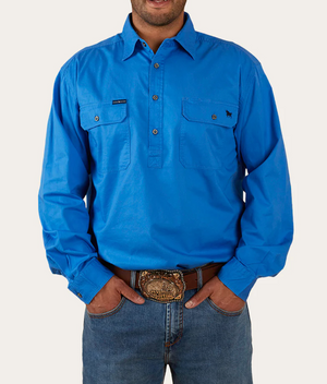 A person is wearing a Ringers Western King River Half Button Shirt in bright blue with two chest pockets and long sleeves, paired with dark jeans. The 100% cotton shirt has a small embroidered logo on one pocket. They stand with hands in pockets, highlighting a detailed belt buckle.