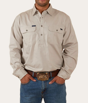 A person is wearing a Ringers Western King River Half Button Shirt in beige. The long-sleeved shirt features two chest pockets, one adorned with an embroidered design. They are also sporting blue jeans and a brown belt with an ornate buckle. The person's hands rest in their pockets, and the shirt collar is open.
