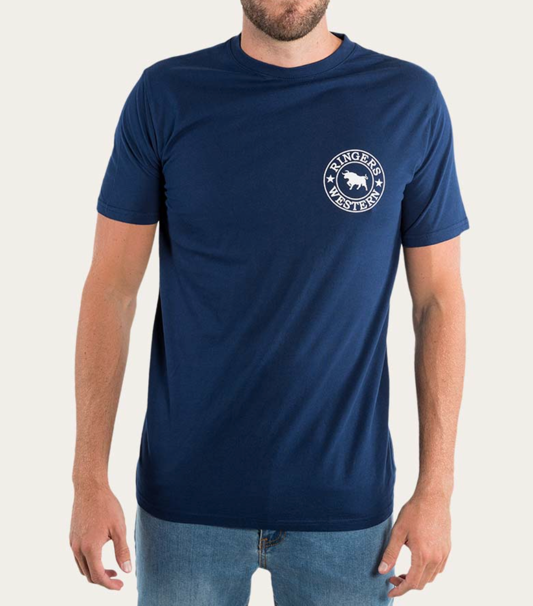 A man wearing a Ringers Western Signature Bull Classic T-Shirt, made from 100% cotton with short sleeves and featuring their logo—a white bull's head in a circular emblem—on the left chest. He pairs the blue tee with light blue jeans against a plain white background.