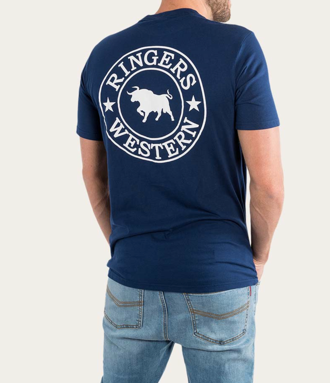 A person stands with their hands behind their back, wearing a navy Ringers Western Signature Bull Classic T-Shirt made from 100% cotton. The shirt showcases the iconic circular logo featuring a bull on the back. They pair it with light blue jeans against a plain white background.