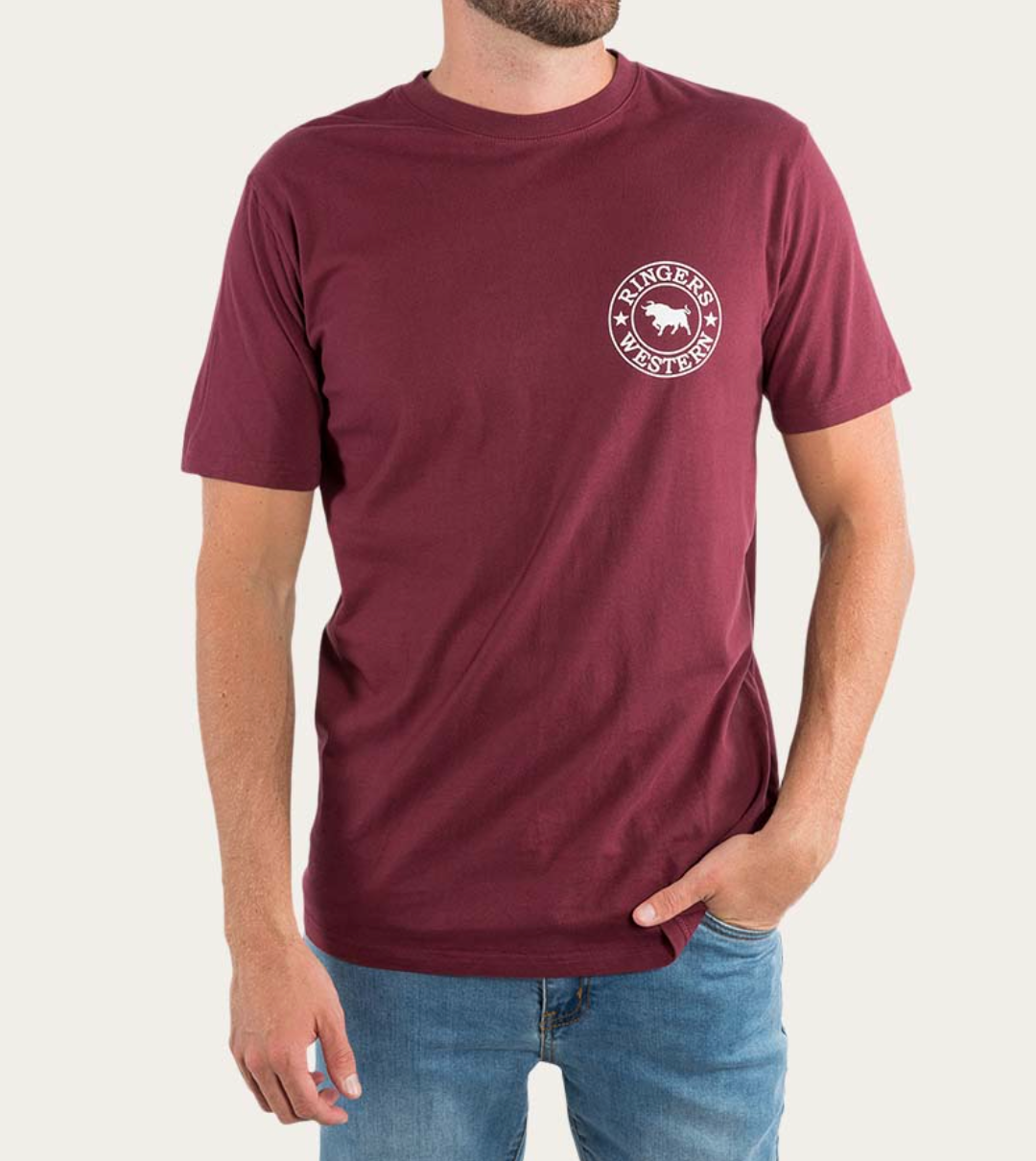 A person wearing the Ringers Western Signature Bull Classic T-Shirt, made of 100% cotton, featuring a maroon short-sleeve design with a circular white logo on the left chest that reads "Ringers Western" and showcases a bull image in the center. The person is also wearing blue jeans, has one hand in their pocket, and is standing against a plain background.