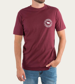 A person wearing the Ringers Western Signature Bull Classic T-Shirt, made of 100% cotton, featuring a maroon short-sleeve design with a circular white logo on the left chest that reads "Ringers Western" and showcases a bull image in the center. The person is also wearing blue jeans, has one hand in their pocket, and is standing against a plain background.