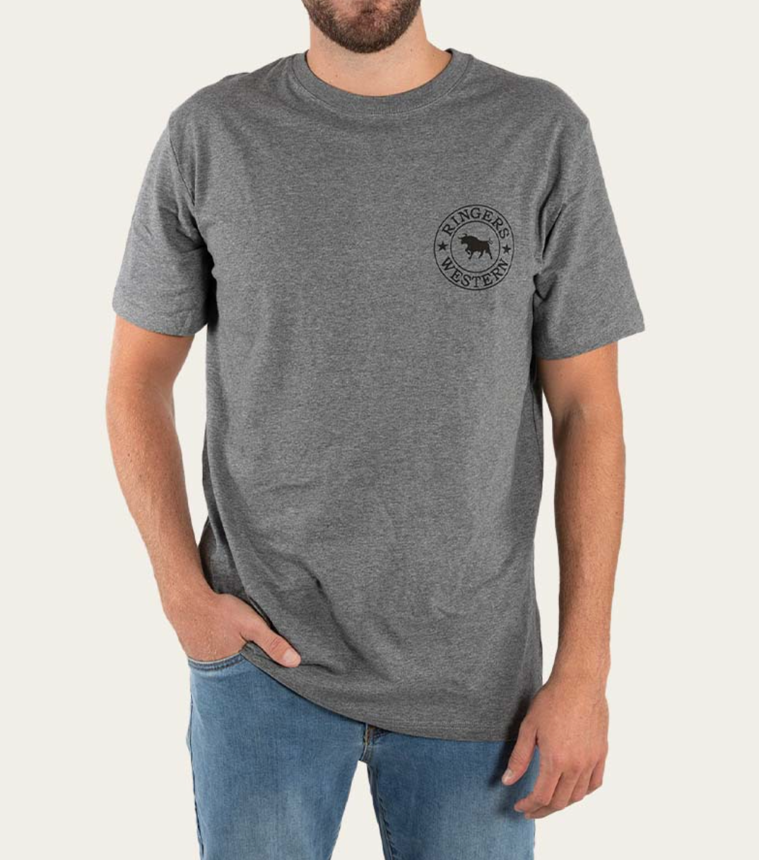 Wearing a Ringers Western Signature Bull Classic T-Shirt made of 100% cotton with a chest logo, a person pairs it with blue jeans, standing against a plain backdrop with one hand in their pocket.