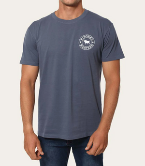 A person dons a Ringers Western Signature Bull Classic T-Shirt in blue, showcasing a circular logo with text and a bull illustration on the upper left side, paired with blue jeans. The classic-style tee is made from pre-shrunk cotton against an off-white backdrop.