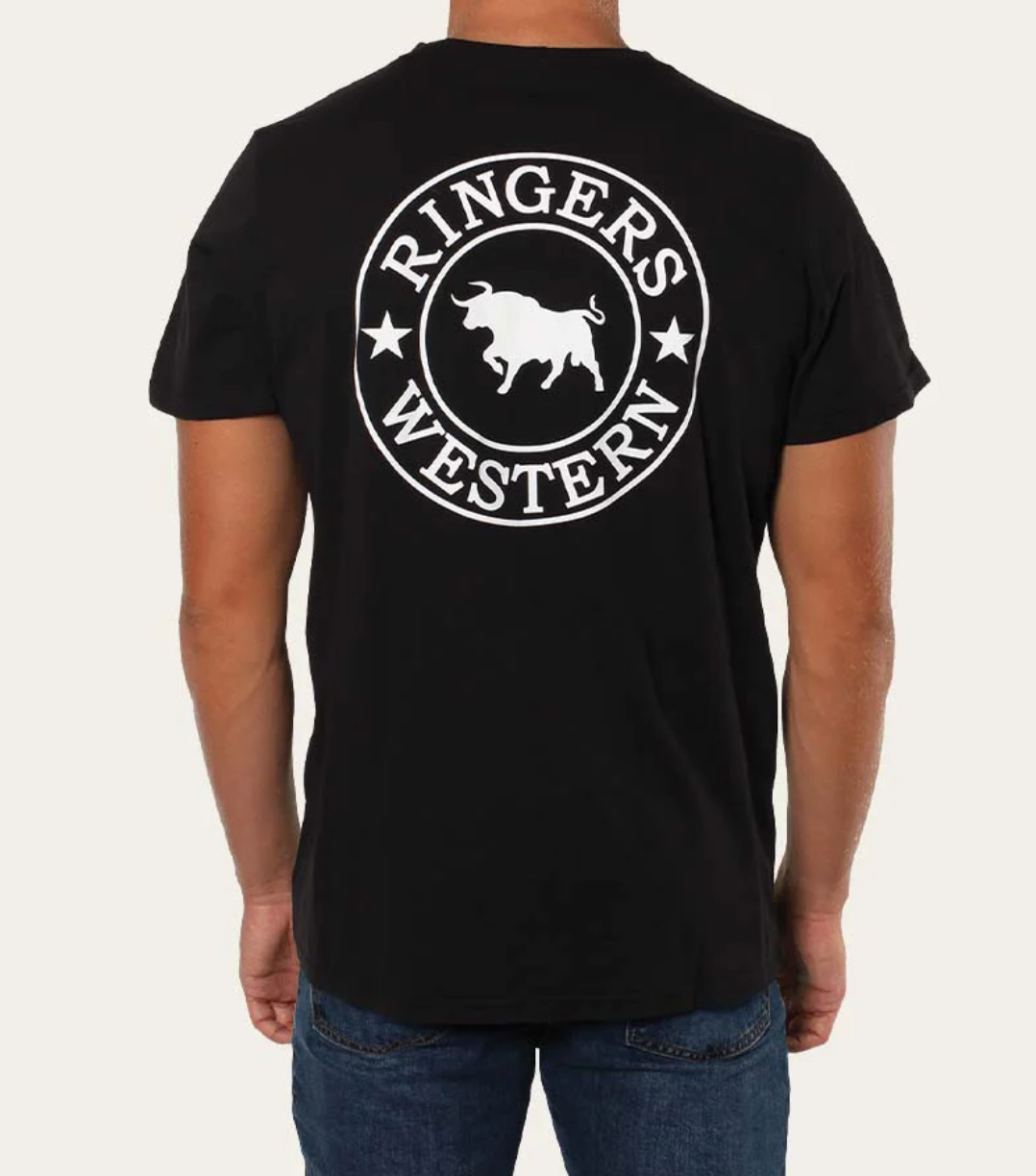 A person is wearing the Ringers Western Signature Bull Classic T-Shirt, a comfortable black tee made of pre-shrunk cotton. The back features a striking white circular logo with a central bull flanked by stars and the words "Ringers Western" encircling it. They pair it with blue jeans and are facing away from the camera.