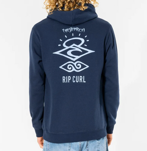A person with curly hair is wearing a Rip Curl Search Icon Hood, featuring "The Search" and abstract symbols on the back. Made from Organic Brushed Cotton, it includes fleece lining for comfort. They're also dressed in light blue jeans.