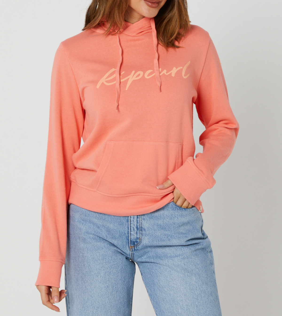A person in a comfy coral Rip Curl Script II Hood with "Ripcurl" on it and light blue jeans stands against a white background. The casual hoodie includes a front pocket where the person's hand rests.