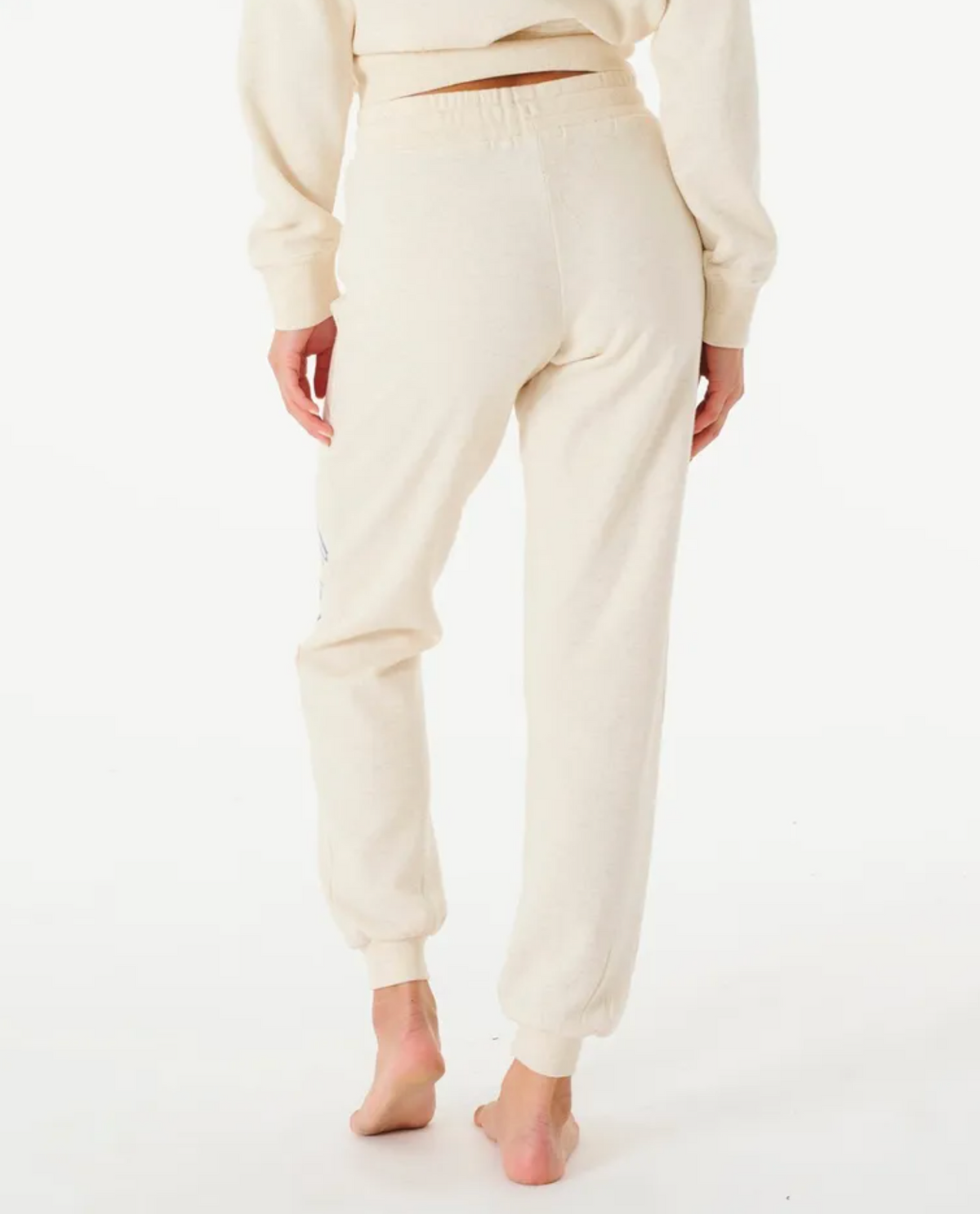 A barefoot person on a white background wears the Rip Curl Script Pant II, a cream-colored sweatshirt, and matching trackpants with an elastic waist, shown from the back focusing on their lower half.