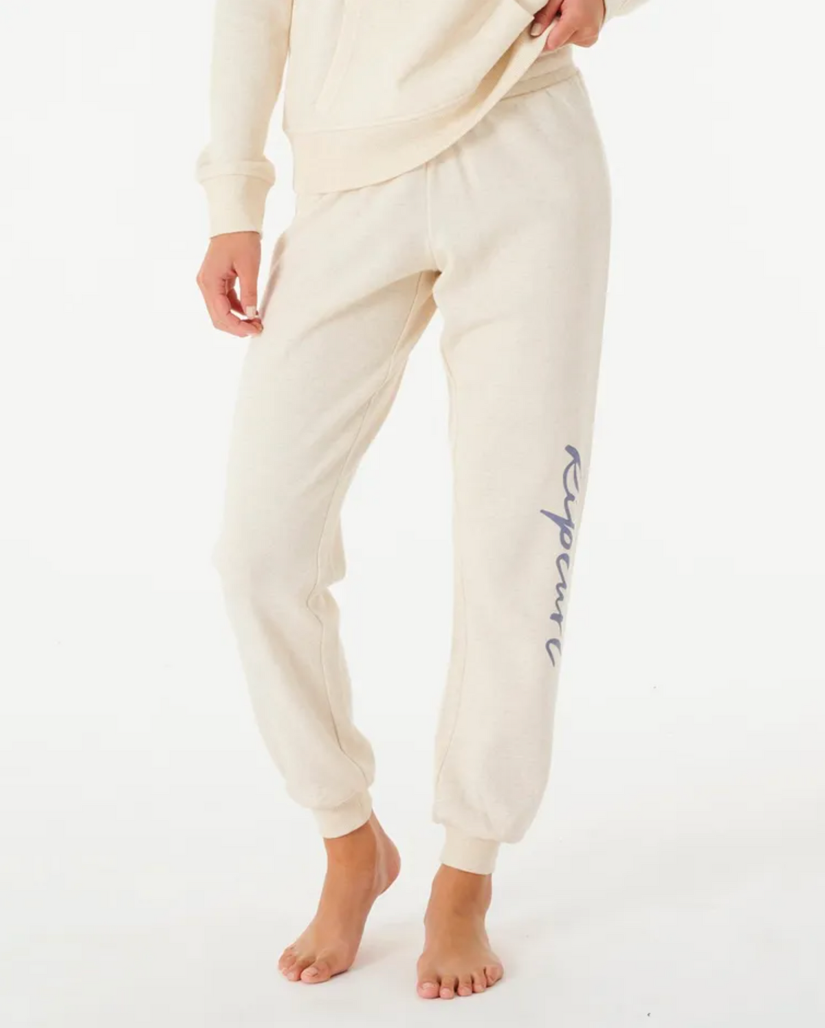 Barefoot on a white background, someone wears Rip Curl Script Pant II in cream, featuring an elastic waist and blue script on the left leg. The comfortable cotton/polyester blend material showcases the lower half of their body.