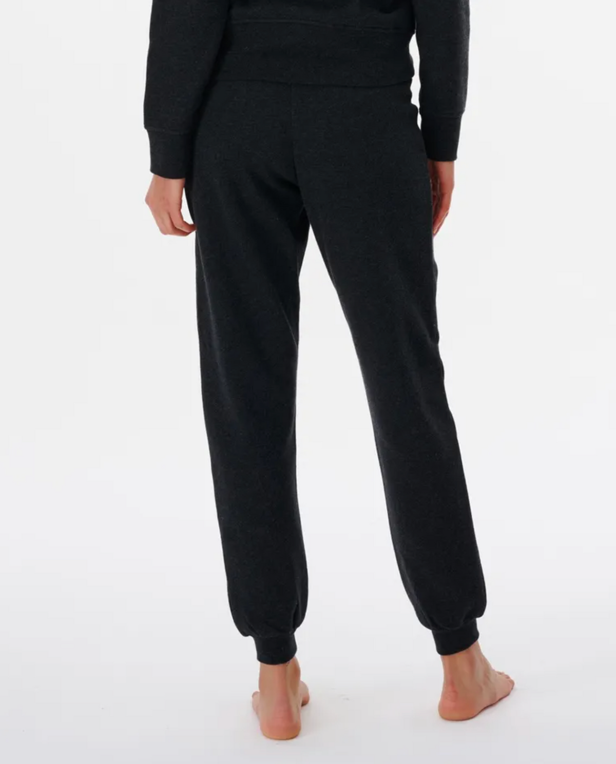 On a white background, someone stands barefoot in dark gray joggers and a matching top. The Rip Curl Script Pant II, crafted from a soft Cotton/Polyester blend with an elastic waist, softly embraces their form as they face away from the camera with hands casually at their sides.