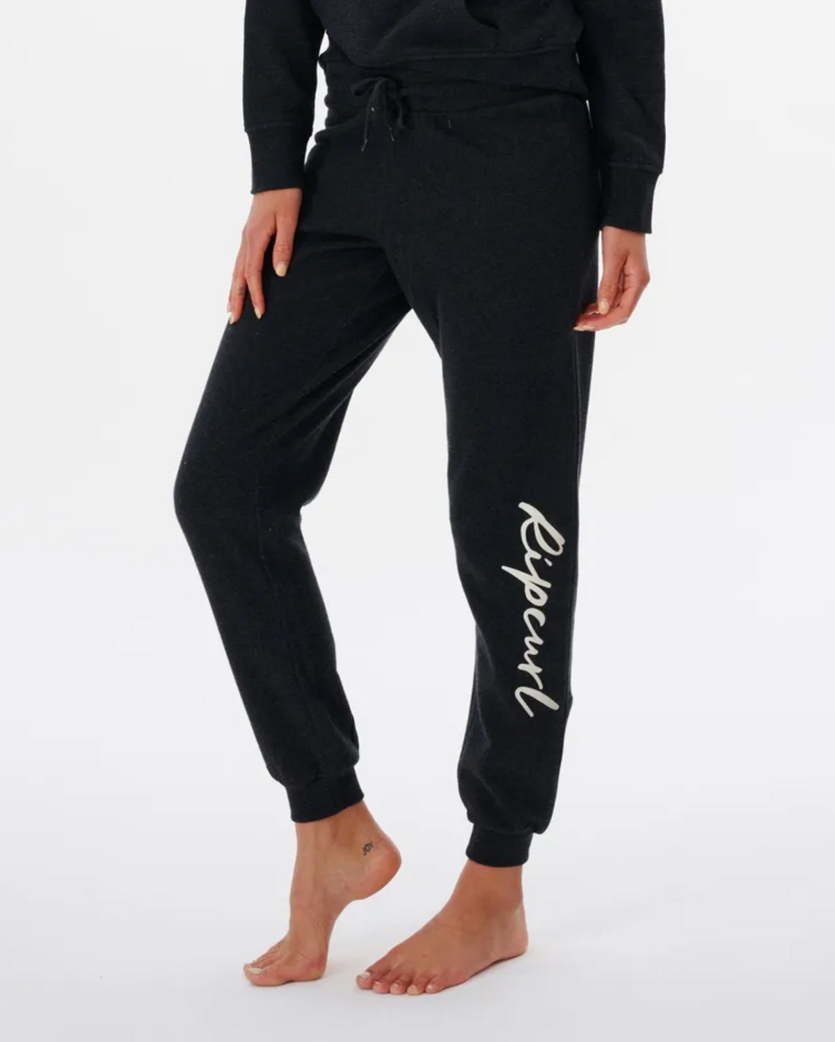 A person is wearing Rip Curl Script Pant II trackpants, featuring a drawstring and elastic waist with "Rip Curl" on the left leg. Made from a Cotton/Polyester blend, they're paired with a matching dark top and seen barefoot against a plain white backdrop.