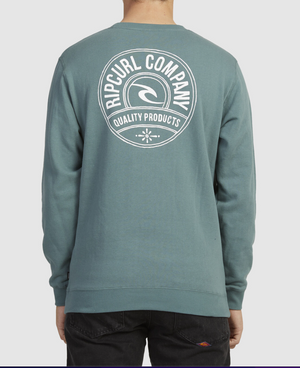 Someone in a teal Rip Curl Stapler Crew sweatshirt, featuring the "Rip Curl Company Quality Products" logo in white on the back, stands facing away against a plain gray background, paired with black pants.