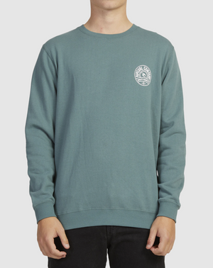 A person is seen in a teal Rip Curl Stapler Crew sweatshirt featuring a small circular logo with text on the left chest, paired with black pants. The head isn't visible, and the background is plain white.