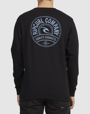 A person wears a Rip Curl Stapler Crew sweatshirt, black with a cotton crew neck. On the back is a blue circular logo reading "Rip Curl Company" and "Quality Products," as they stand facing away, hands in pockets.