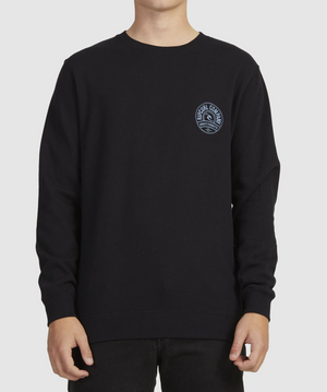 A person wears the Rip Curl Stapler Crew, a black crew neck sweatshirt with a small round logo featuring text and a wave graphic on the upper left side, paired with black cotton pants. Their head is not visible in the image.