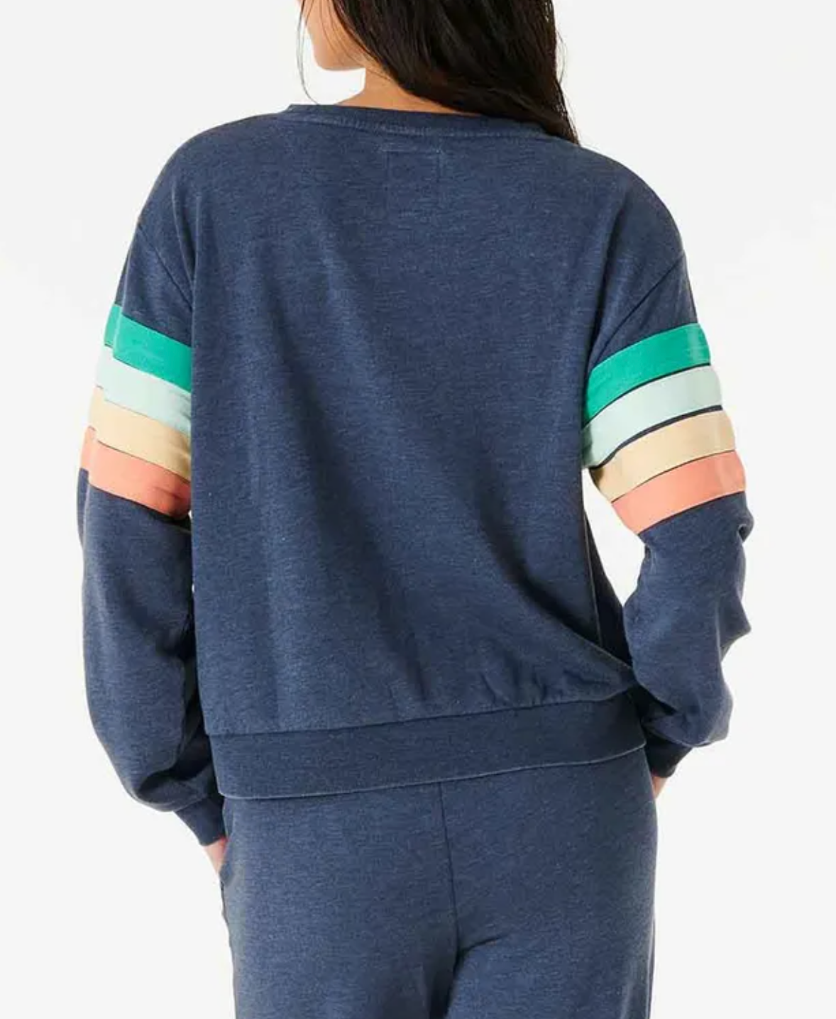 A person with long hair is wearing the Rip Curl Day Break Crew, a relaxed fit blue sweatshirt and pants. The crew neck sweatshirt features colorful sleeve stripes in green, turquoise, peach, and beige. The person is facing away from the camera.