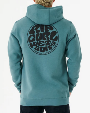 A person with curly hair is wearing a blue Rip Curl Wetsuit Icon Hood, showcasing a bold circular logo on the back that reads "Rip Curl Wet Suits, Torquay, Trestles, Hossegor." Made from organic cotton, the hoodie blends style and sustainability as they face away from the camera.