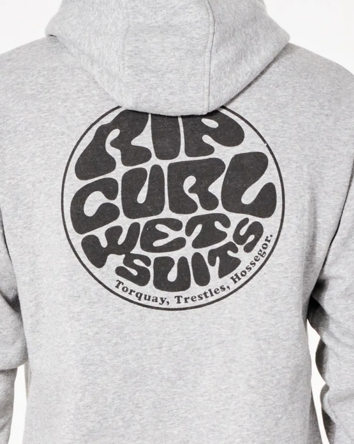 A person is shown from the back wearing a gray Rip Curl Wetsuit Icon Hood. The cozy hoodie has a prominent circular logo with the text "Rip Curl Wetsuits" in a retro, wavy font. Below the logo, three locations are listed: Torquay, Trestles, Hossegor.