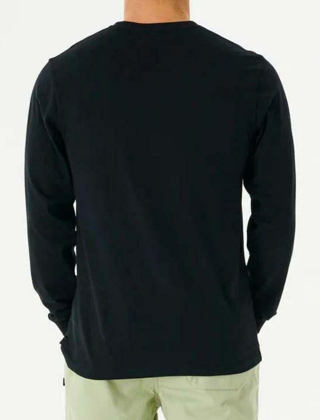 The image depicts a person wearing a Rip Curl Fade Out Icon Long Sleeve Tee, which is black and made of organic cotton, paired with light-colored pants. They are seen from behind against a plain white background.