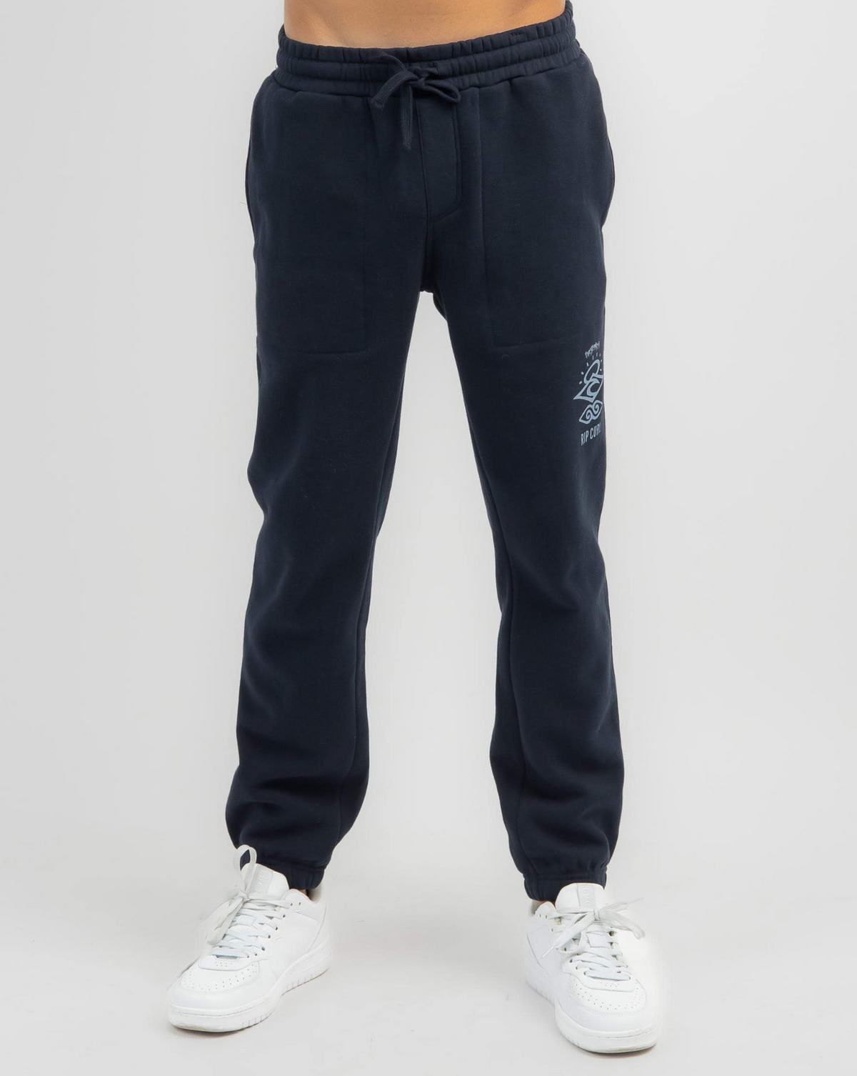 A person wearing dark blue Rip Curl Search Icon Track Pants made from organic brushed cotton with a drawstring waist and small logo on the side stands against a plain background, paired with white sneakers.