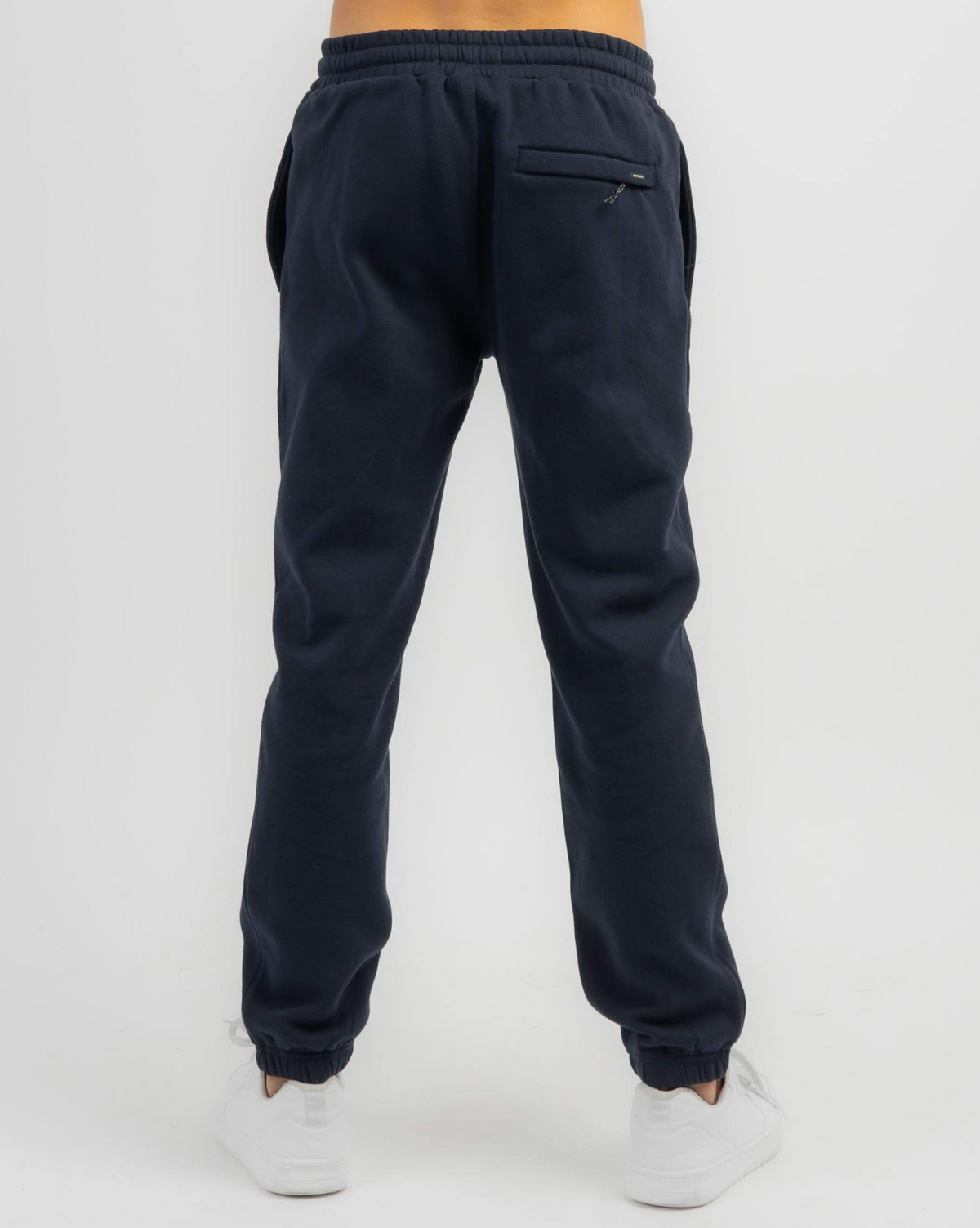 Back view of a person in Rip Curl Search Icon Track Pant, made from organic brushed cotton, with zippered back pocket, elastic waistband and cuffs. Paired with white sneakers on a plain white background, highlighting this stylish and eco-friendly choice.