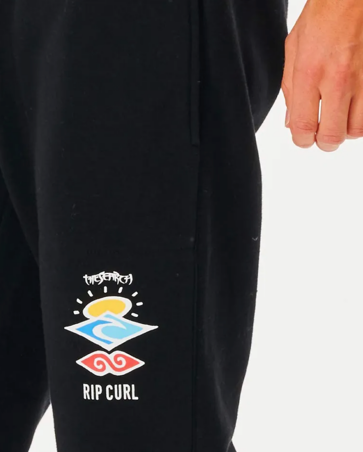 A close-up of someone in black Rip Curl Search Icon Track Pants, adorned with colorful sun, wave, and spiral graphics. "Rip Curl" is visible below the designs. Made from organic brushed cotton, their hand rests casually by their side.