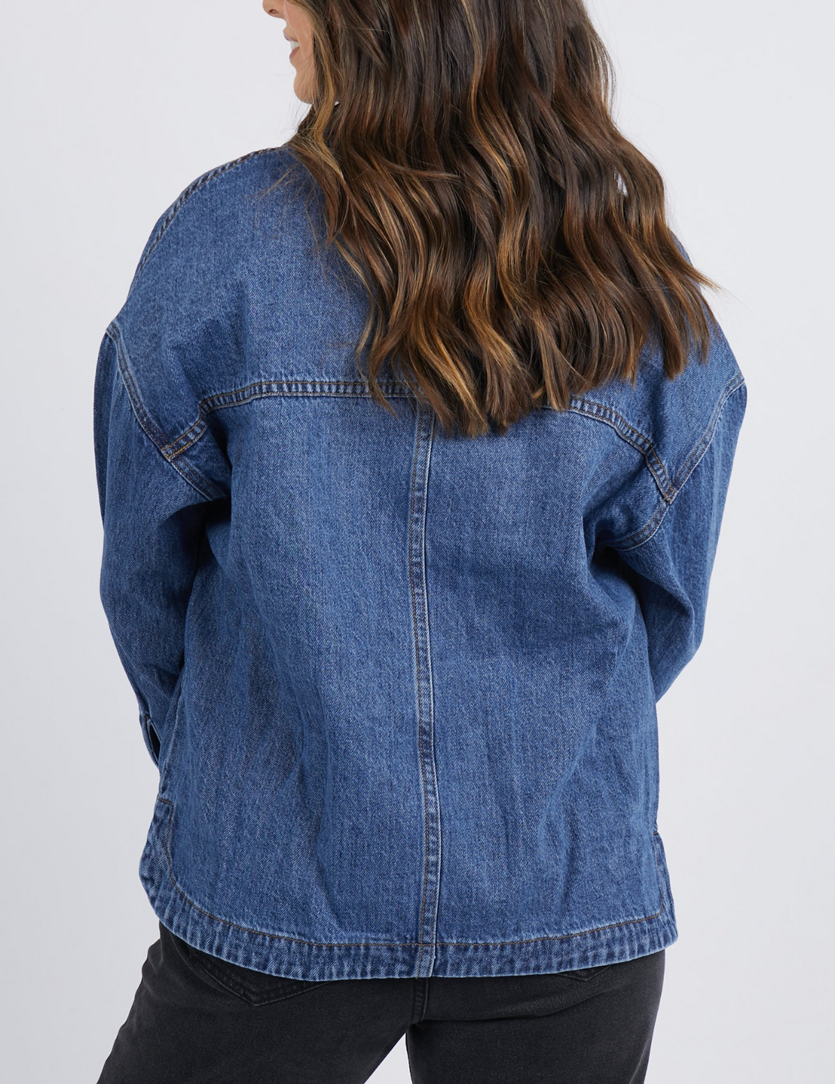A woman with wavy brown hair, facing away, wears the Foxwood Joelle Denim Shacket and black pants against a gray background. The relaxed fit adds comfort and style while showcasing the shacket's 100% cotton material.