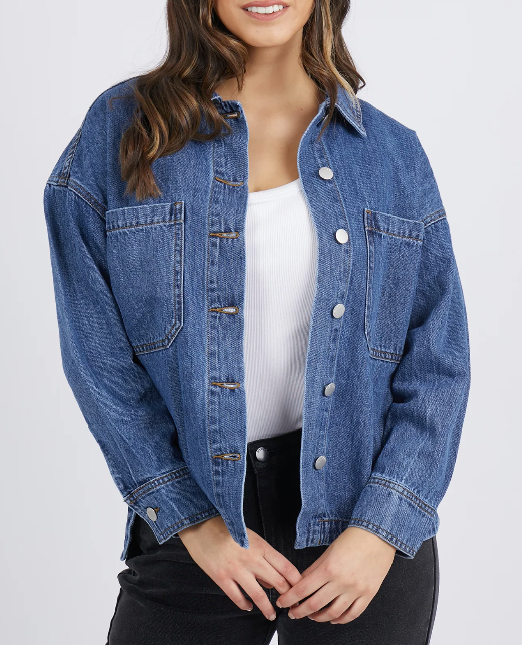 A person stands against a plain, light background with loose, wavy hair. They wear the Foxwood Joelle Denim Shacket, a relaxed fit 100% cotton blue denim piece with front pockets and metal buttons, layered over a white shirt and paired with black pants.