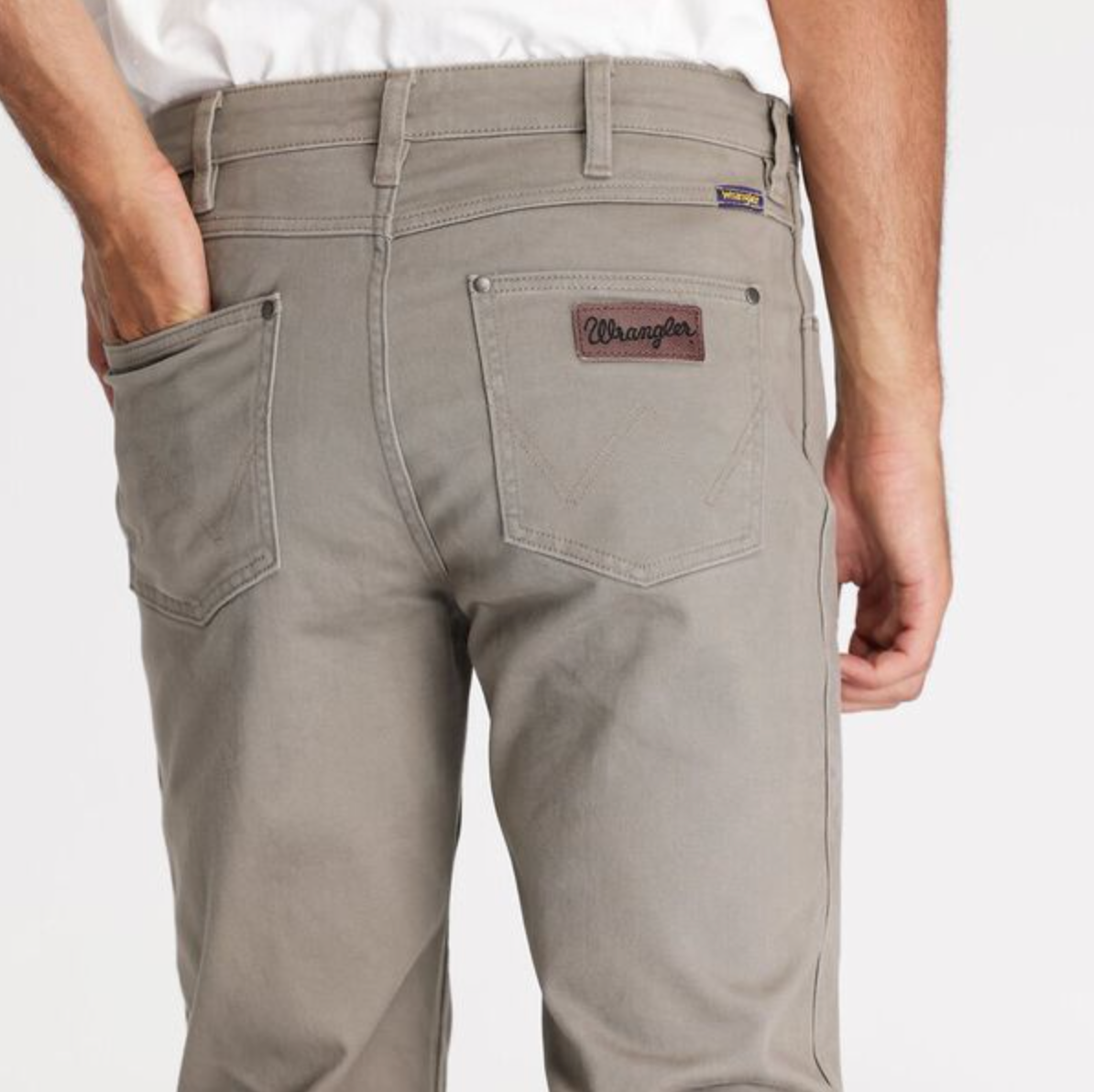 Dressed in a white shirt, someone showcases Wrangler Classics Straight Moleskin Jeans, featuring water-saving technology. The gray jeans have a logo patch above the right pocket, casually resting under their hand.