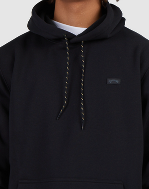 Someone is shown from shoulders to waist in a black Billabong Shoreline Pullover hoodie with yellow drawstrings and a small rectangular chest patch, set against a light background, emphasizing the pullover's heavyweight fabric.